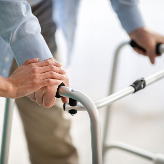 Unrecognizable elderly man using walker, young doctor supporting and helping him at retirement home. Pensioner moving around with walking frame, caregiver holding his hand, closeup